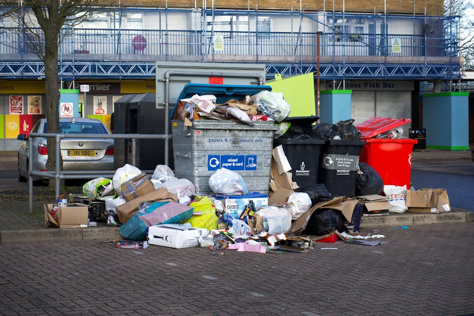 A large pile of mixed waste and rubbish is accumulated on a paved pedestrian area, adjacent to a parking space with a car showing a yellow registration plate. The waste includes various black and blac