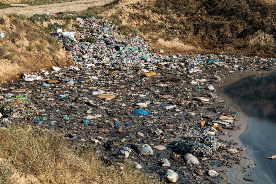 A large pile of mixed waste and rubbish is accumulated on a paved pedestrian area, adjacent to a parking space with a car showing a yellow registration plate. The waste includes various black and blac